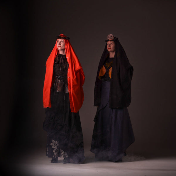 Two models standing with head covering and in long layered clothing. Dark moody light 