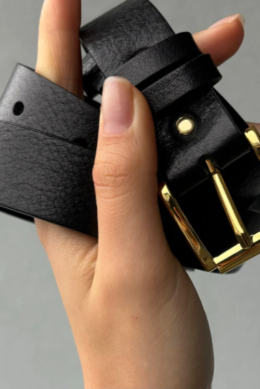 Black leather belt with gold buckle held by a hand against a gray background